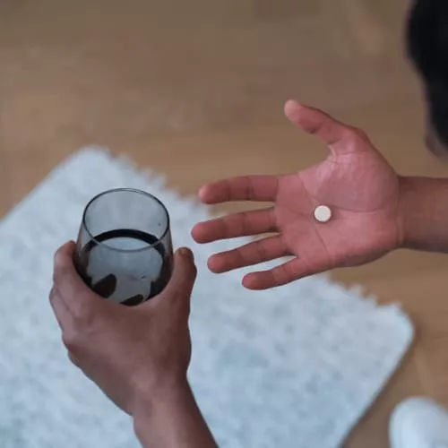 A man's hands, one holding a white tablet, the other holding a glass of water.